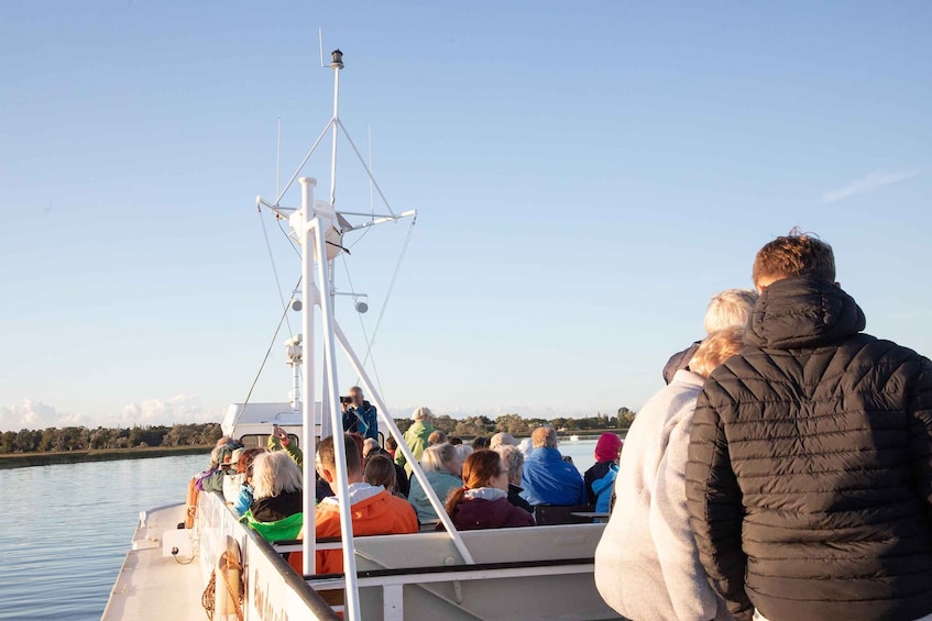 A group of people on a boat, facing away from the camera, with a calm body of water and a clear sky in the background. 1
