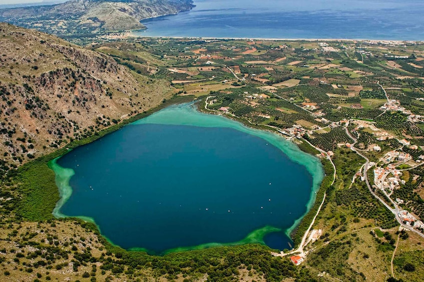 An aerial view of a coastal area with a large turquoise lake surrounded by hilly terrain and scattered buildings. 1