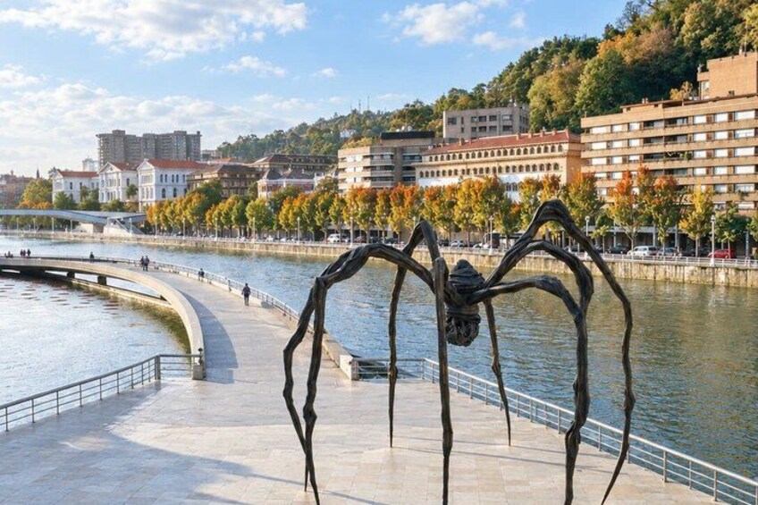 A waterfront promenade with a large metal sculpture of a spider, a curved walkway, and buildings in the background. 4