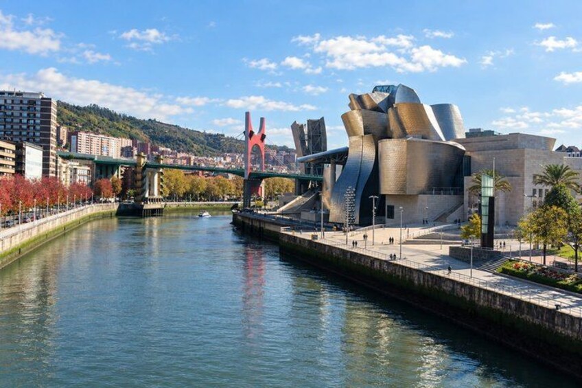 A modern building with a unique, reflective facade by the river, a red bridge, and surrounding urban landscape. 2