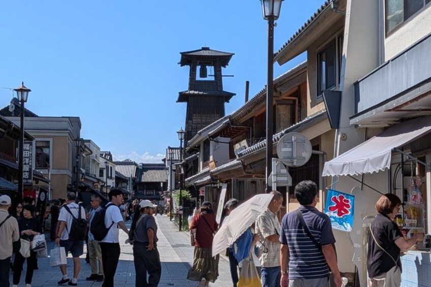 A busy street with people walking, traditional buildings, and a tower in the background. 2