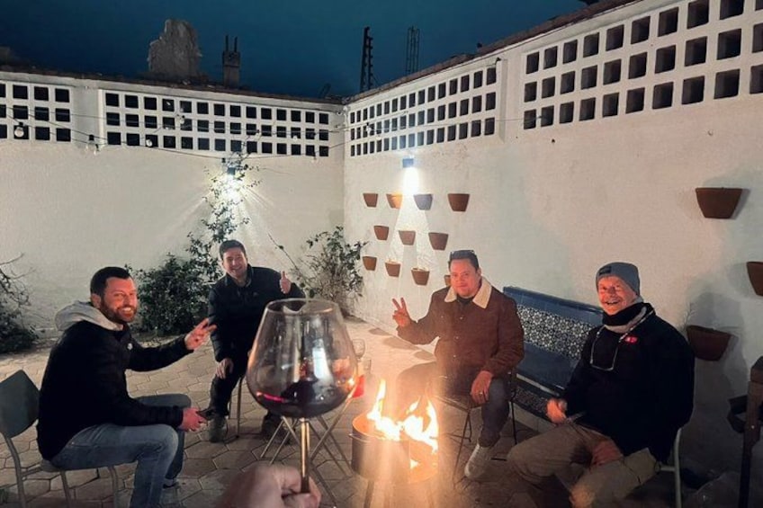 A group of people sitting around a fire with a glass of wine, in an outdoor setting with a white wall and potted plants. 1