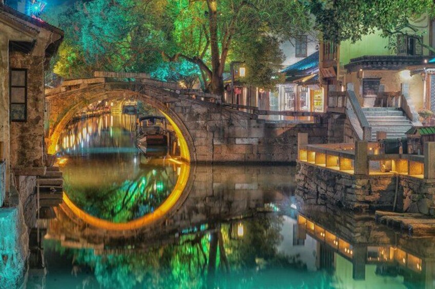A stone bridge over a calm water body, illuminated by warm lights, with traditional buildings and trees in the background. 2