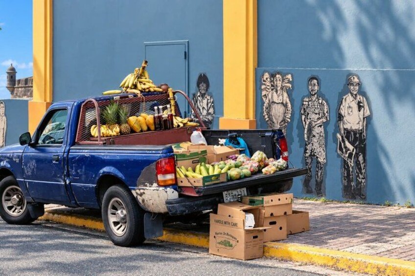 A blue pickup truck loaded with fruits and boxes parked in front of a mural. 3