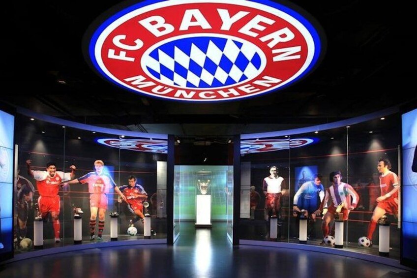 A sports hall with a large FC Bayern Munich logo, trophies, and players' statues. 4