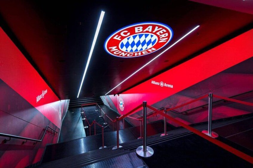 An indoor area with a prominent FC Bayern Munich logo, red and black walls, and a staircase leading upwards. 1