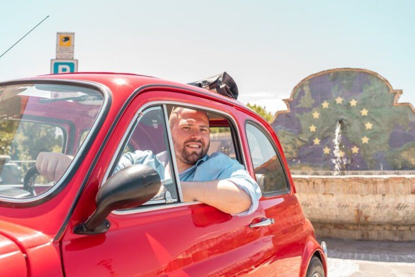 A red car parked near a fountain with a European Union flag backdrop. 1