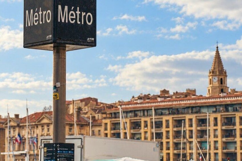 A metro sign with the word 'Metro' in both English and Spanish, a clock tower, and a building with a red roof in the background. 5