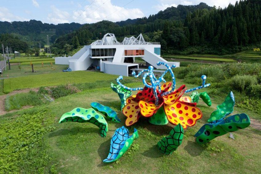 A colorful sculpture of a flower and insects in a grassy area with a modern building in the background. 1