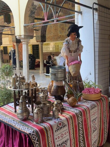 A person in traditional attire standing by a table with various metal containers and a large pot, in a courtyard with arches and a tiled wall. 2