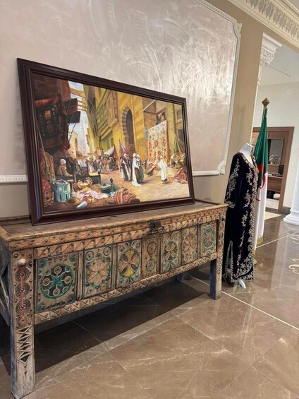 A framed painting of a historical scene with people and a market, displayed on a decorated cabinet. 3