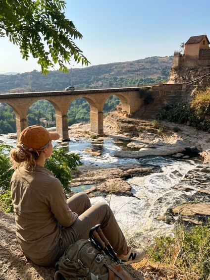 A person sitting by a river with a stone bridge in the background. 1