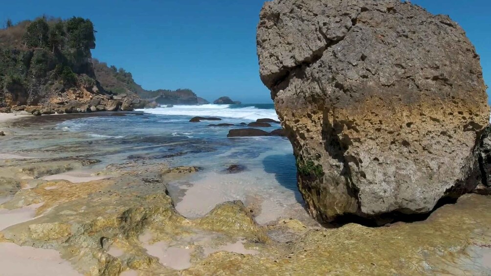 A rocky beach with clear shallow waters and a large boulder in the foreground. 3