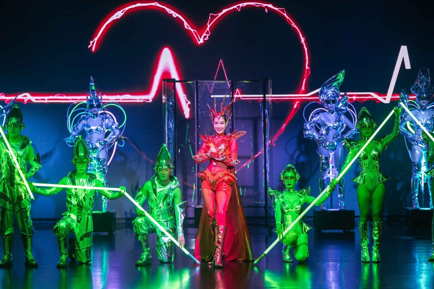 Eine Gruppe von Darstellern in futuristischer Kleidung mit Neonlichtern vor einem neonbeleuchteten Herzschild. 1