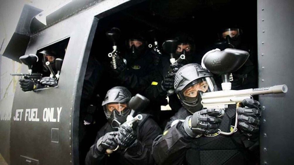 A group of people in black tactical gear with helmets and masks, holding paintball guns, inside a vehicle marked 'JET FUEL ONLY'. 2