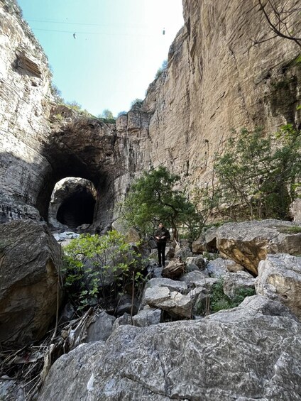 A person standing in a rocky canyon with a large cave opening. 5