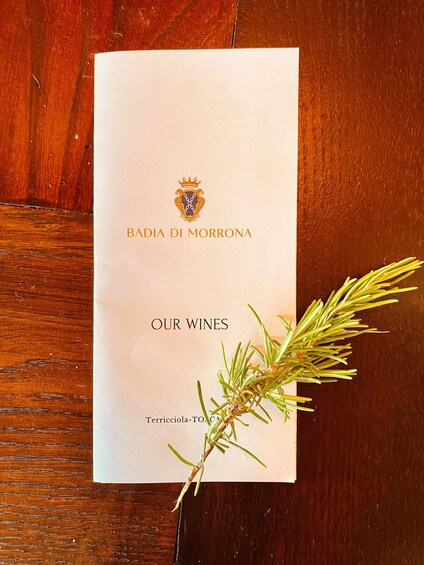 A menu titled 'BADIA DI MORRONA OUR WINES' with a sprig of rosemary on a wooden table. 2