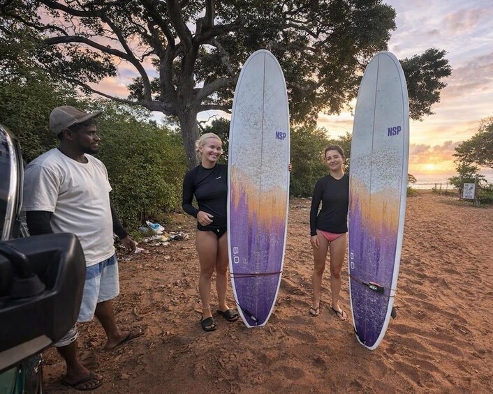Drei Personen tragen Surfbretter an einem sandigen Strand, im Hintergrund Bäume und ein Sonnenuntergang. 2