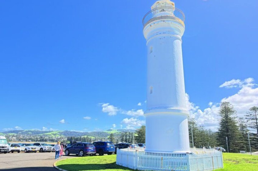 A tall white lighthouse with a metal railing, surrounded by a fenced area, with cars and a person in the background. 3