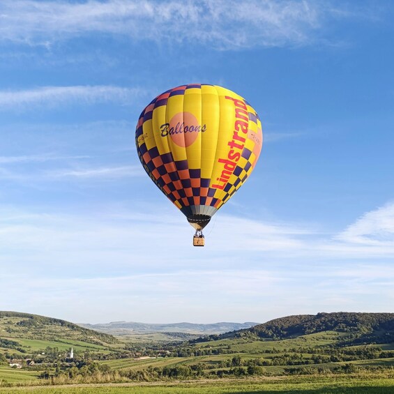 Une montgolfière ornée des inscriptions « Ballons » et « Lindstrass », survolant un paysage verdoyant avec des collines et une église blanche. 4