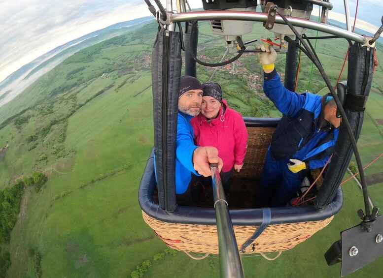 Une nacelle de montgolfière avec deux personnes à l’intérieur, l’une portant une veste bleue et l’autre une veste rose, survolant un paysage verdoyant. 3