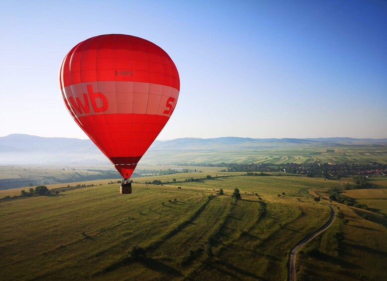 Une montgolfière rouge vif, portant les inscriptions « C-OEFS » et « WbS », survole un paysage rural composé de champs et d’un petit village. 1