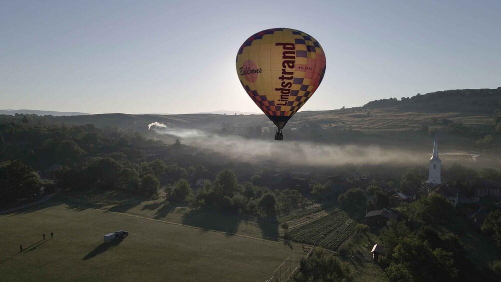 Un ballon à air chaud portant l’inscription « Lindstrand » survolant un paysage rural avec des champs, une église et une route. 2