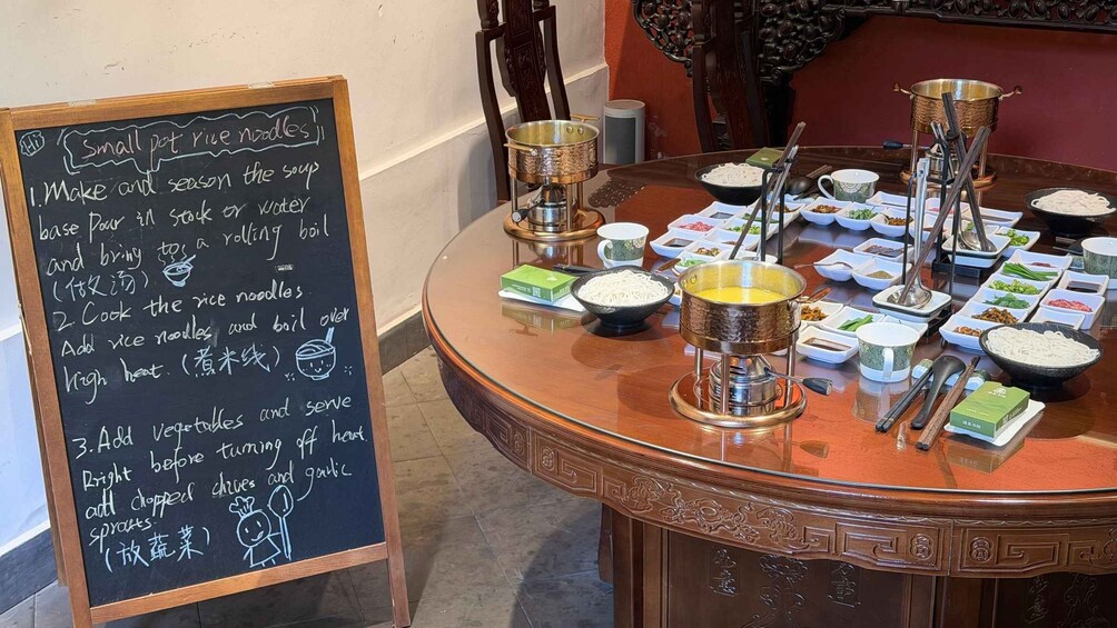 Un tableau noir sur lequel sont écrites des instructions de cuisine, une table ronde avec différents plats et ustensiles de cuisine. 1