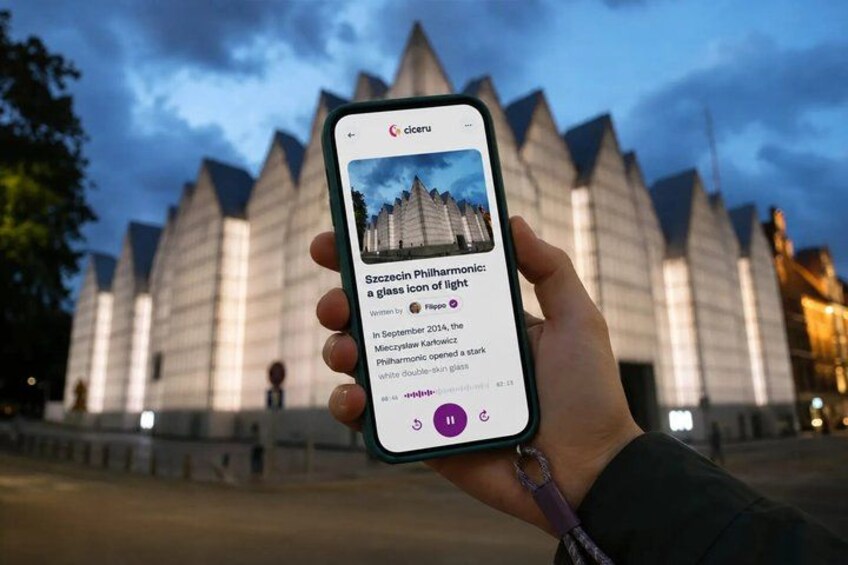A hand holding a smartphone displaying an article titled 'Szczecin Philharmonic: a glass icon of light' with a background of a modern building with pointed roofs. 1