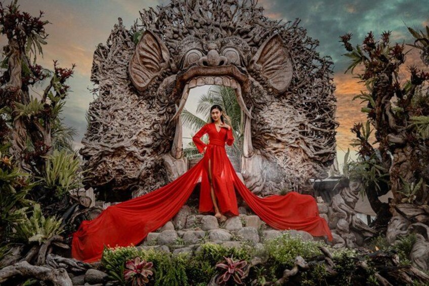 A person in a red dress standing in front of a large, ornate archway surrounded by tropical plants and rocks. 1