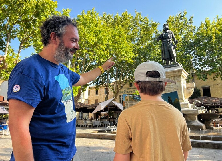 Un homme désigne une statue sur une place publique, avec des arbres et des tables en plein air en arrière-plan. 5
