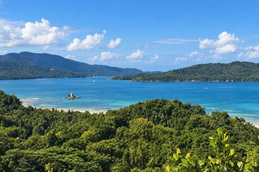 A coastal landscape with a clear blue sea, dense green forest, and distant mountains under a partly cloudy sky. 2