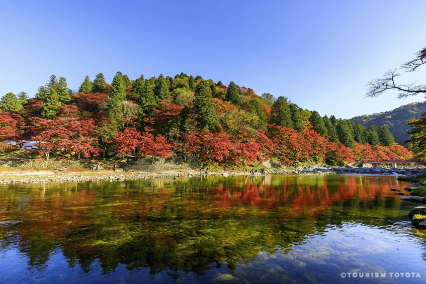 Un lago sereno con reflejos de follaje rojo y verde, rodeado por un promontorio boscoso y un cielo azul despejado. 3