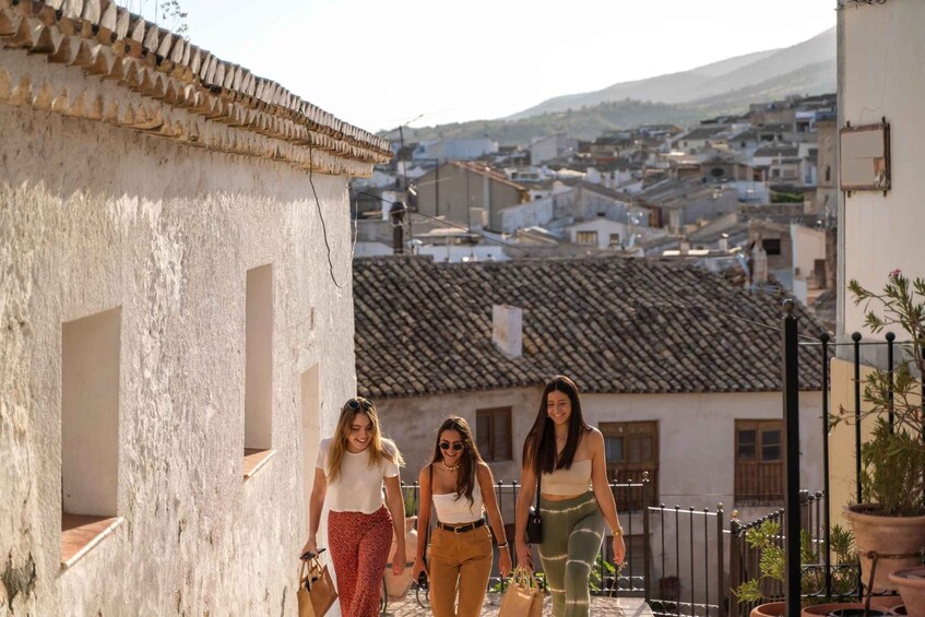 Drei Frauen gehen auf einer Straße entlang, mit traditioneller Bebauung und einer Bergkulisse im Hintergrund. 1
