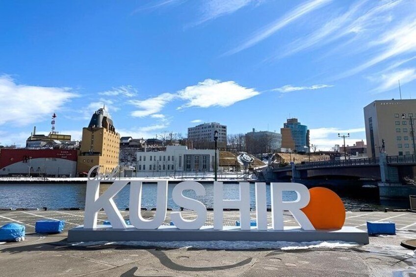 A large sign with the word 'KUSHIR' and a circle, set against a cityscape with buildings and a river. 2