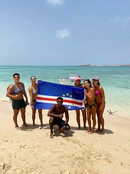 Eine Gruppe von Menschen am Strand, eine Flagge, klares blaues Wasser und ein Boot im Hintergrund. 2