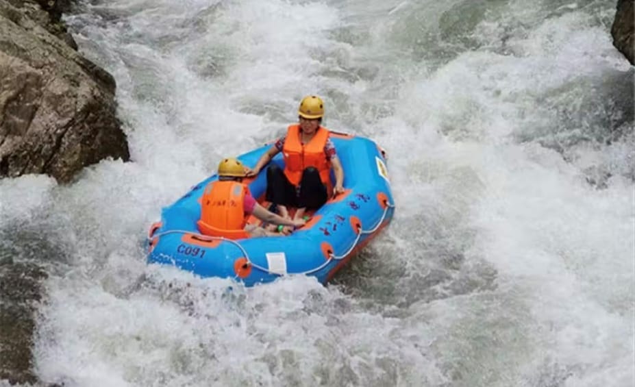 A blue inflatable raft with two people wearing life jackets and helmets navigating through a river with white water rapids. 1