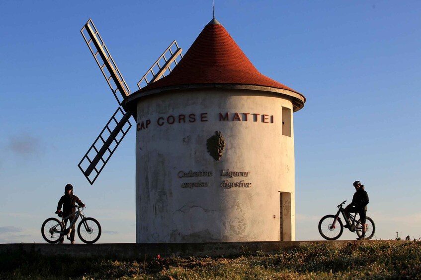 Eine Windmühle mit rotem Dach und einem Schild mit der Aufschrift 'Mattei'. Zwei Radfahrer fahren auf einem Weg davor. 1