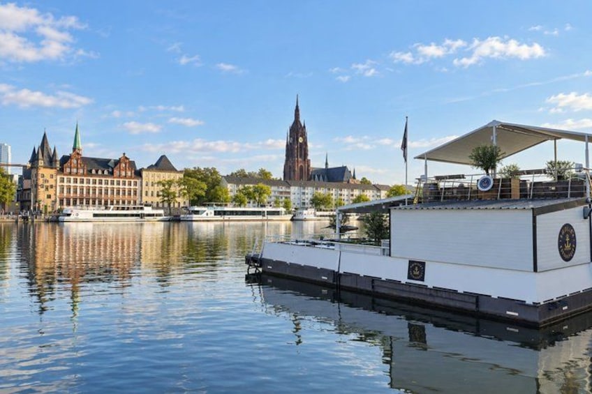 A river with a boat docked on the right, a historic building with a spire in the background, and a clear blue sky. 3