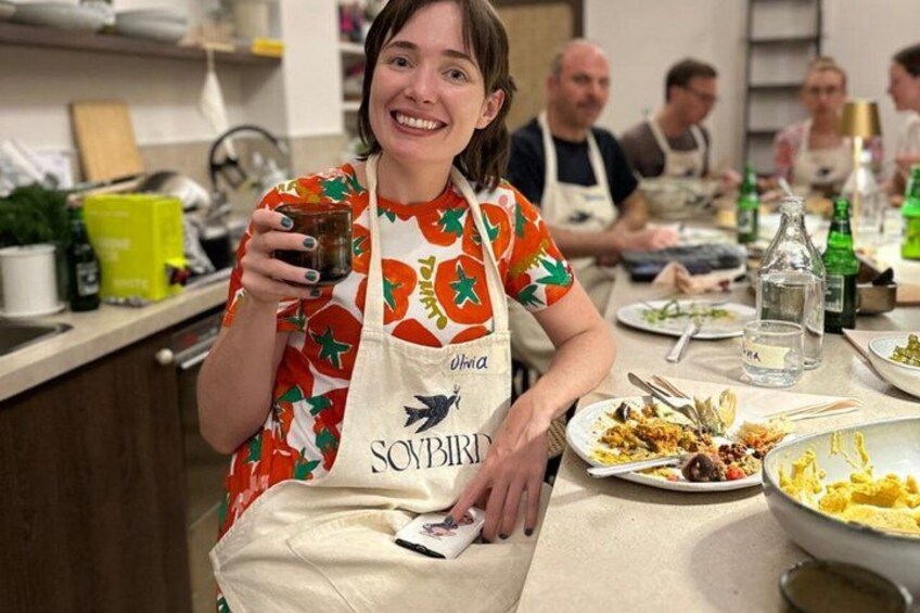 A person holding a jar, wearing a patterned apron, with a table full of dishes and bottles in the background. 4