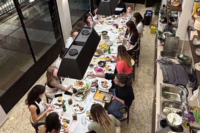 A group of people dining at a long table in a restaurant with a kitchen area visible to the right. 2