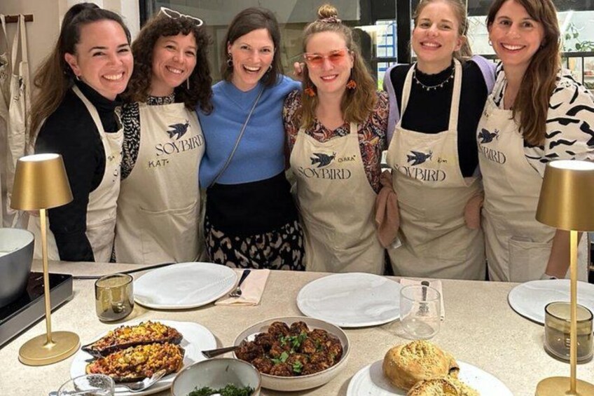A group of people wearing aprons with a logo, standing around a table with various dishes. 3