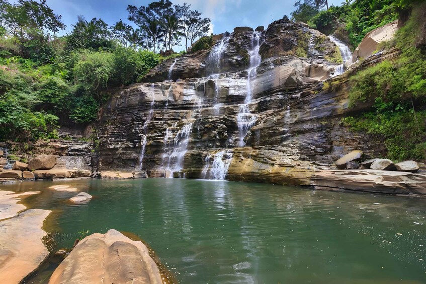 Une cascade d’eau se déversant sur des formations rocheuses en terrasses, pour se jeter dans un bassin d’eau paisible, le tout entouré d’une végétation luxuriante. 2