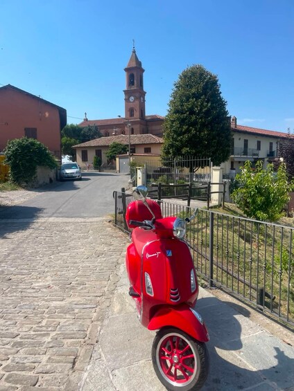 Un scooter Vespa rouge est garé sur une rue pavée, avec une église et des immeubles d’habitation en arrière-plan. 5
