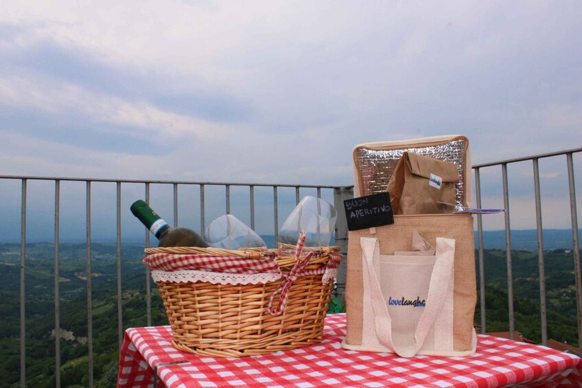 Un coin pique-nique aménagé avec un panier en osier, une bouteille de vin et un sac fourre-tout posés sur une nappe à carreaux, offrant une vue sur un paysage magnifique. 2