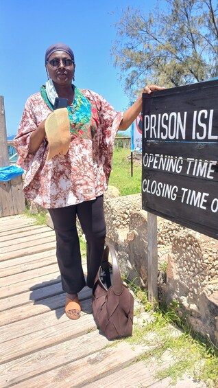 A person standing on a wooden boardwalk holding a sign that reads 'Prison Island' with 'Opening Time' and 'Closing Time' text. 2