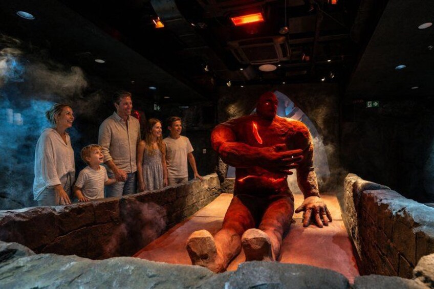 A family observing a large red statue in a dimly lit room with stone walls and a stone platform. 5