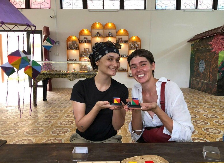 Two people holding Rubik's cubes at a table with patterned floor tiles and a decorative wall with framed pictures. 1