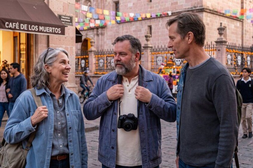 Three people standing on a cobblestone street, a café with a sign, and colorful decorations in the background. 3