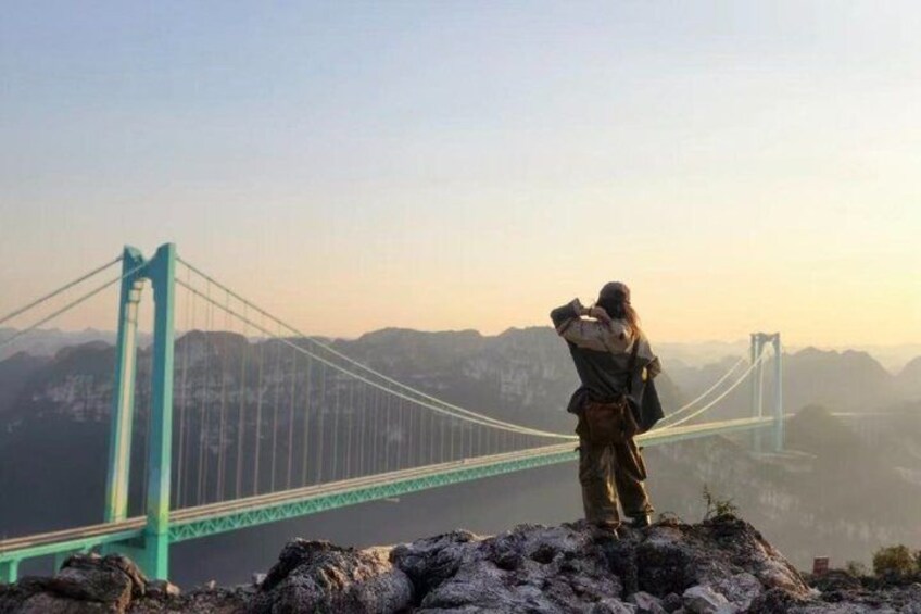 A person standing on a rocky outcrop overlooking a bridge. 1